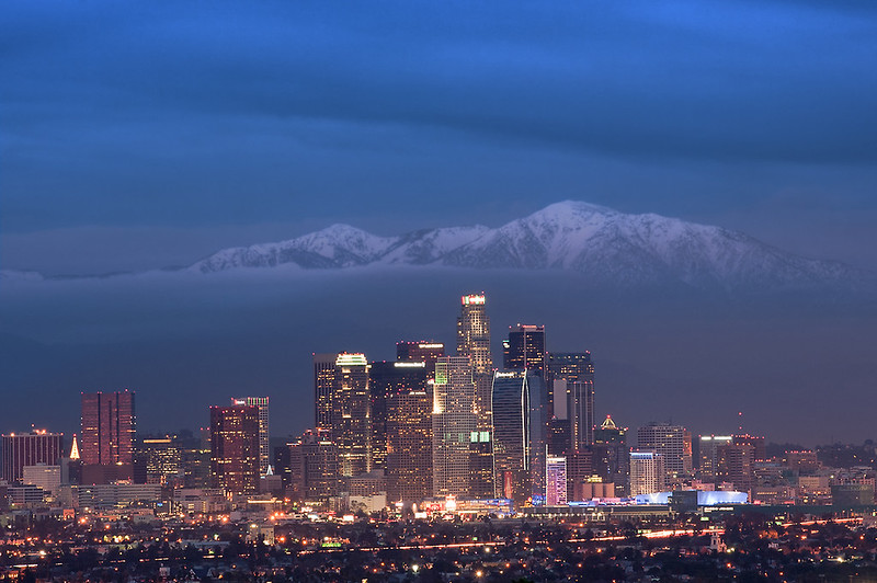 Downtown Los Angeles skyline lit up at night, snow-covered mountains and clouds in the background.