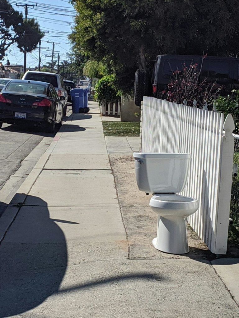suburban sidewalk, white picket fence, cars parked on the side of the road...and a porcelain toilet sitting outside the fence.
