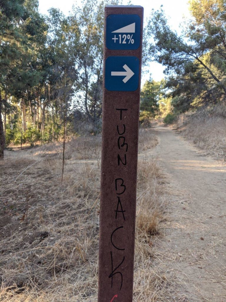 Signpost for a trail in a wooded area, with TURN BACK scrawled on it.