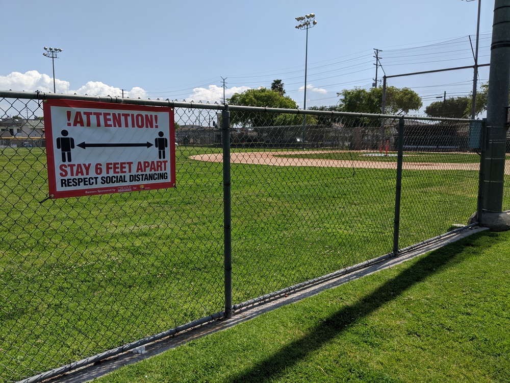 Fence at a park with a baseball field in the distance, and a sign saying ATTENTION! STAY 6 FEET APART. RESPECT SOCIAL DISTANCING.
