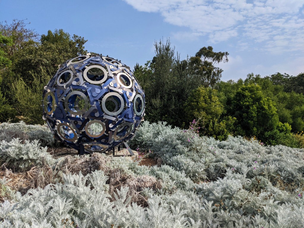 A blue-and-silver lattice sphere outside in a patch of light green ground cover, with brighter green trees and blue sky in the background.