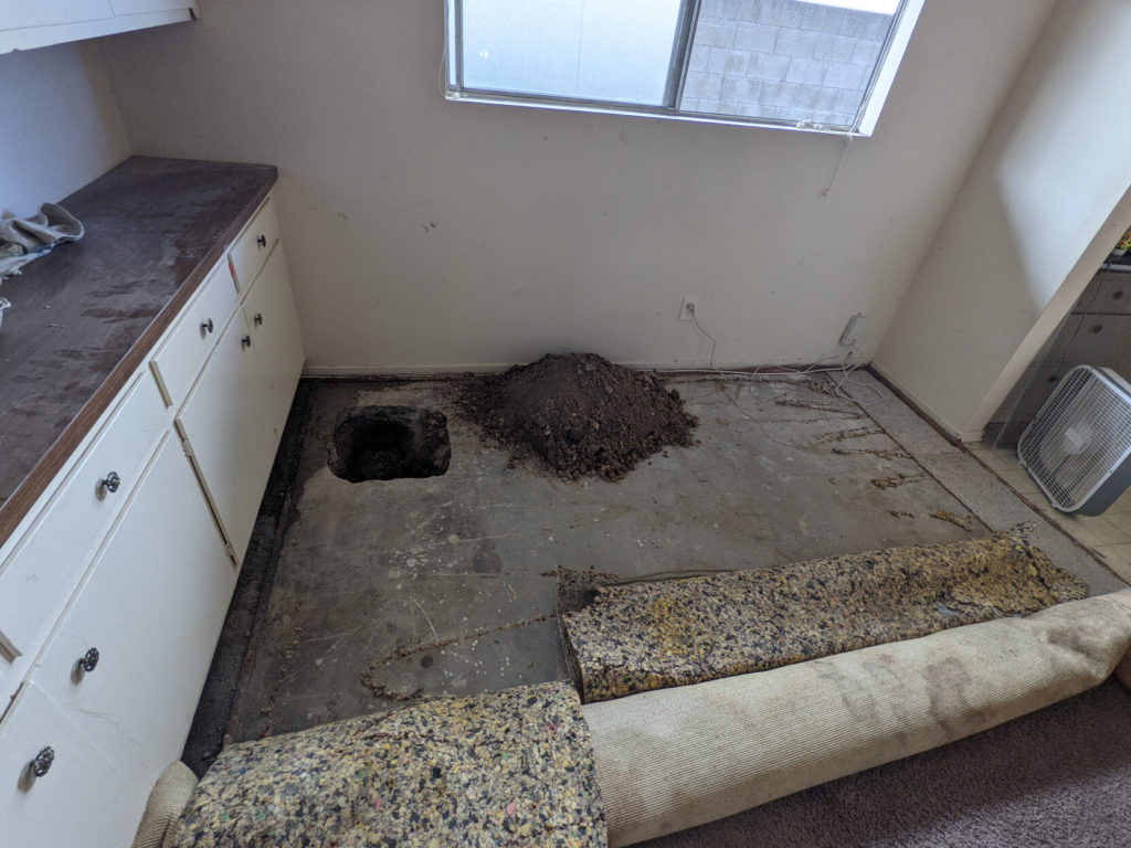 An empty room with the carpet peeled up revealing a concrete floor with a 1.5x1.5 foot hole in the corner and a pile of dirt next to it.