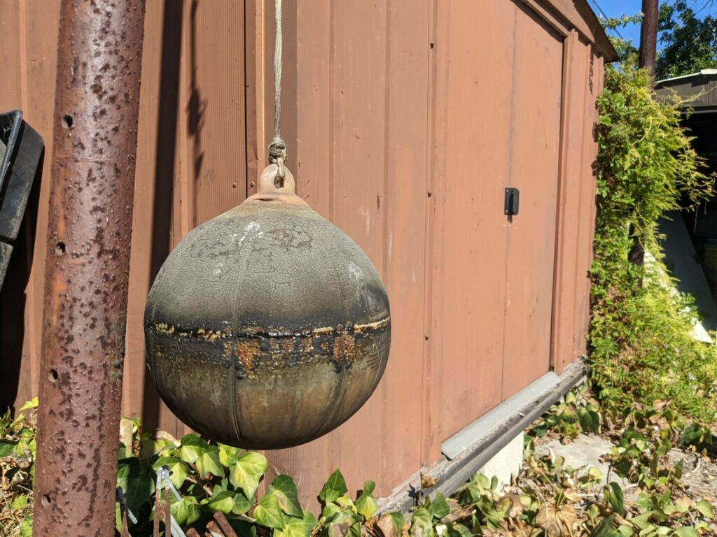 An old tetherball, still hanging from a pole, it's surface darkened and stretched and falling apart with some parts that look shiny as if they're seeping some kind of liquid.