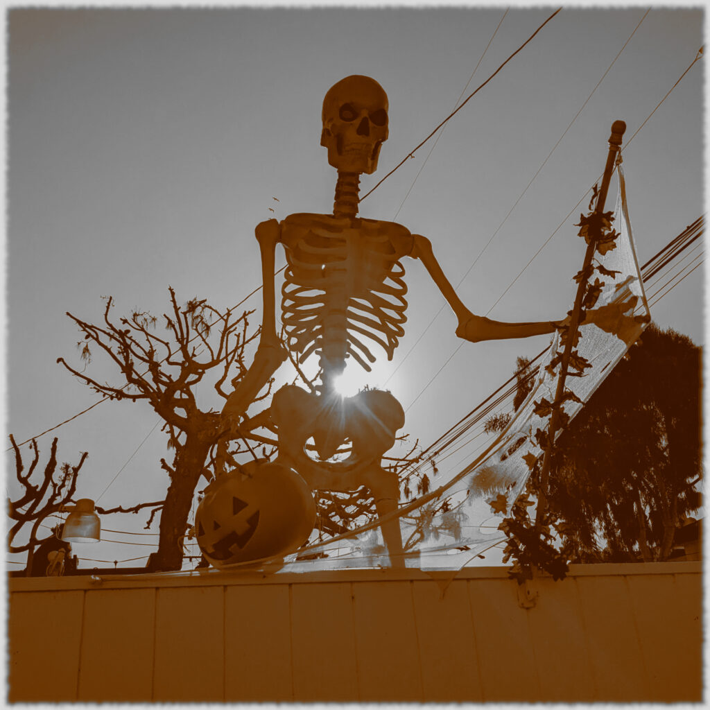 A giant skeleton seen from below, towering over a wall, with the sun directly behind it, all filtered to sepia.