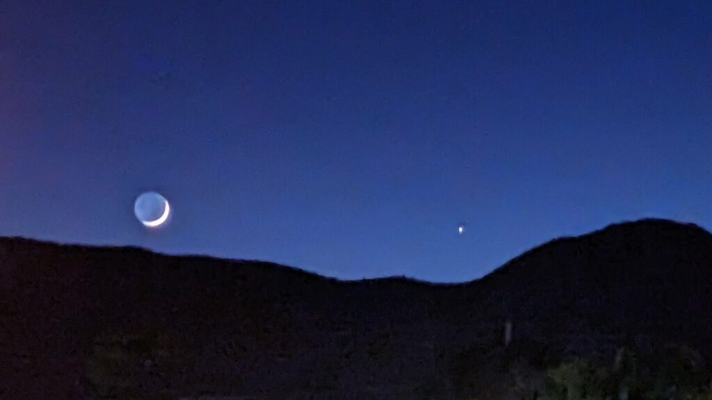 Dark blue sky with silhouetted hills below it. A crescent moon with the dark filled in(but fainter) by Earthshine and a single 'star' are visible just above the hills